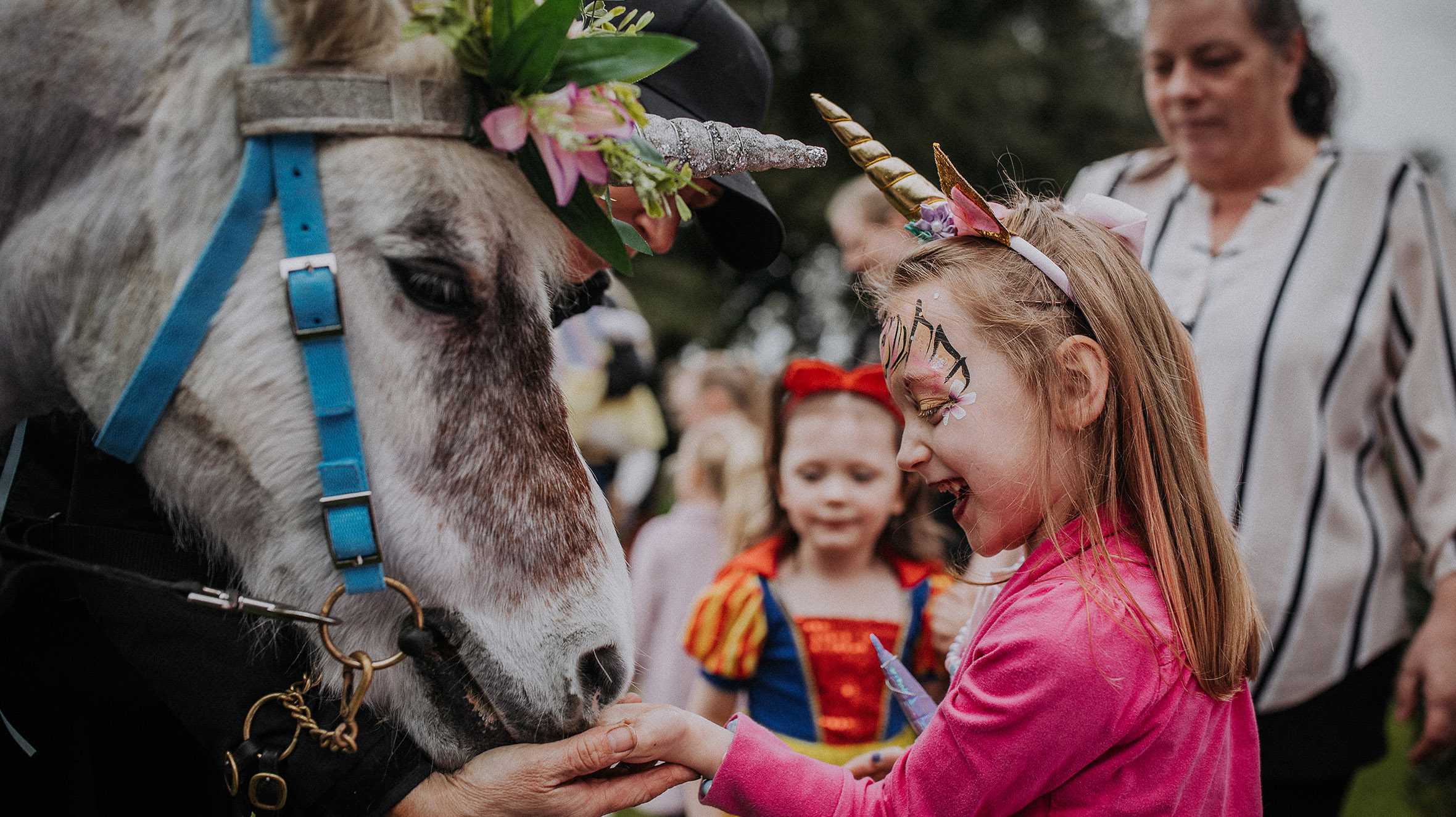 Lacey, wearing a unicorn horn and facepaint, feeds a pony wearing a unicorn horn