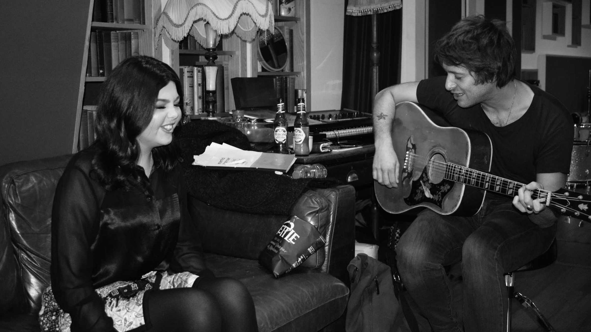 Molly sitting on the sofa smiling, while Paolo Nutini plays the guitar.