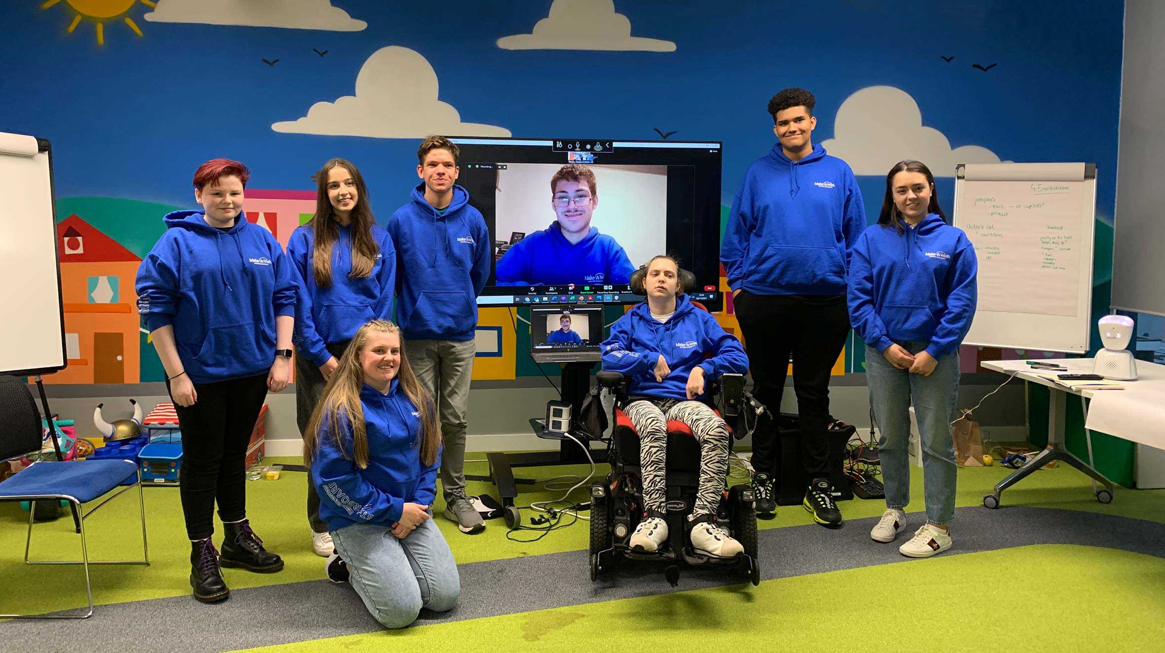 All of the STARboard members standing together in the Make-A-Wish playground, with Josh appearing virtually on a TV screen behind the group.