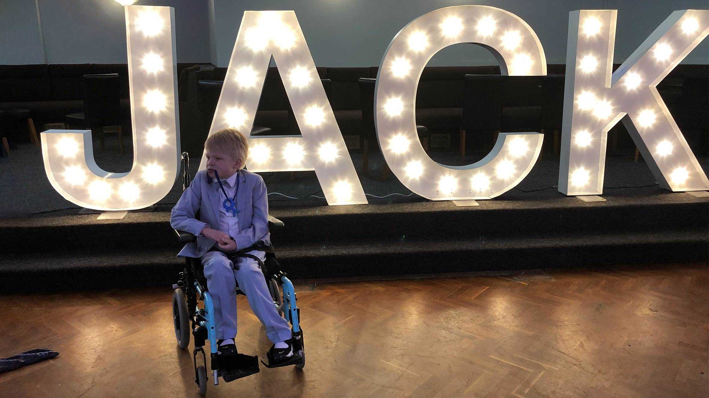 Jack sitting in his wheelchair and wearing a colourful tuxedo, with his name in giant lights behind him.