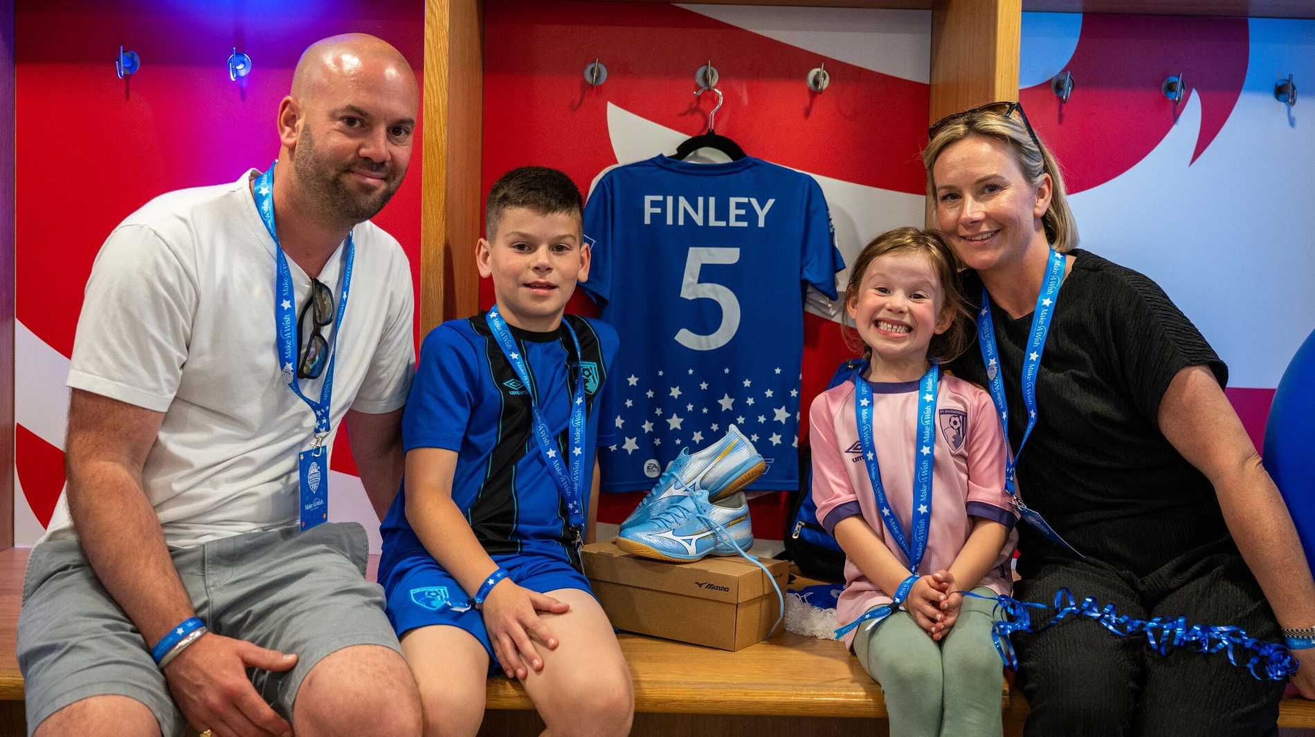 A young boy, Finlay sits proudly in front of his Make-A-Wish Wishes United football shirt, which has his name 'Finlay' and the Number 5 on the back. A man is sat next to him and the other side of his football boots sits a young girl and a lady.