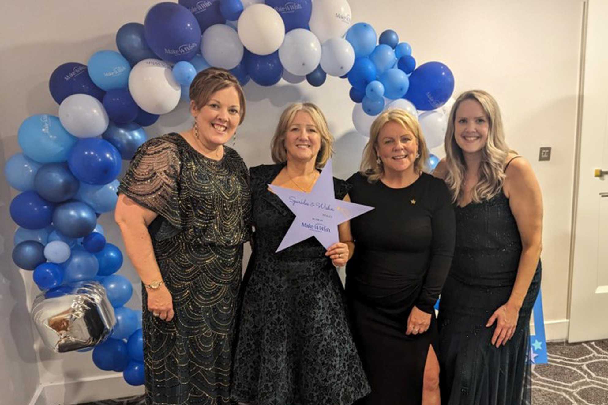 Make-A-Wish volunteers, Sian, Karen, Karen and Rhiannon standing in front of a blue balloon arch in their ball gowns.