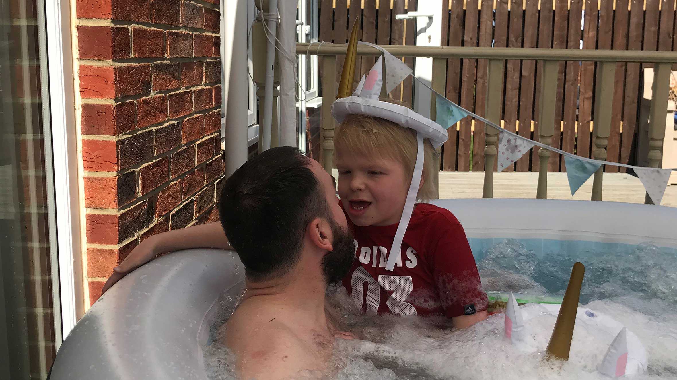 Jack, wearing a red t-shirt and an inflatable unicorn hat, is sitting in a hot tub with his dad and looking into his dad's eyes.