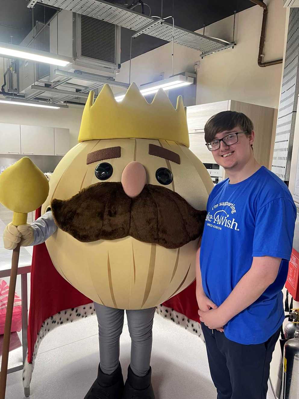 Former Wish Child Alistair stands with his hands together next to the Overcooked character, the Onion King. The Onion King is wearing a crown, has a big moustache and is holding a staff.