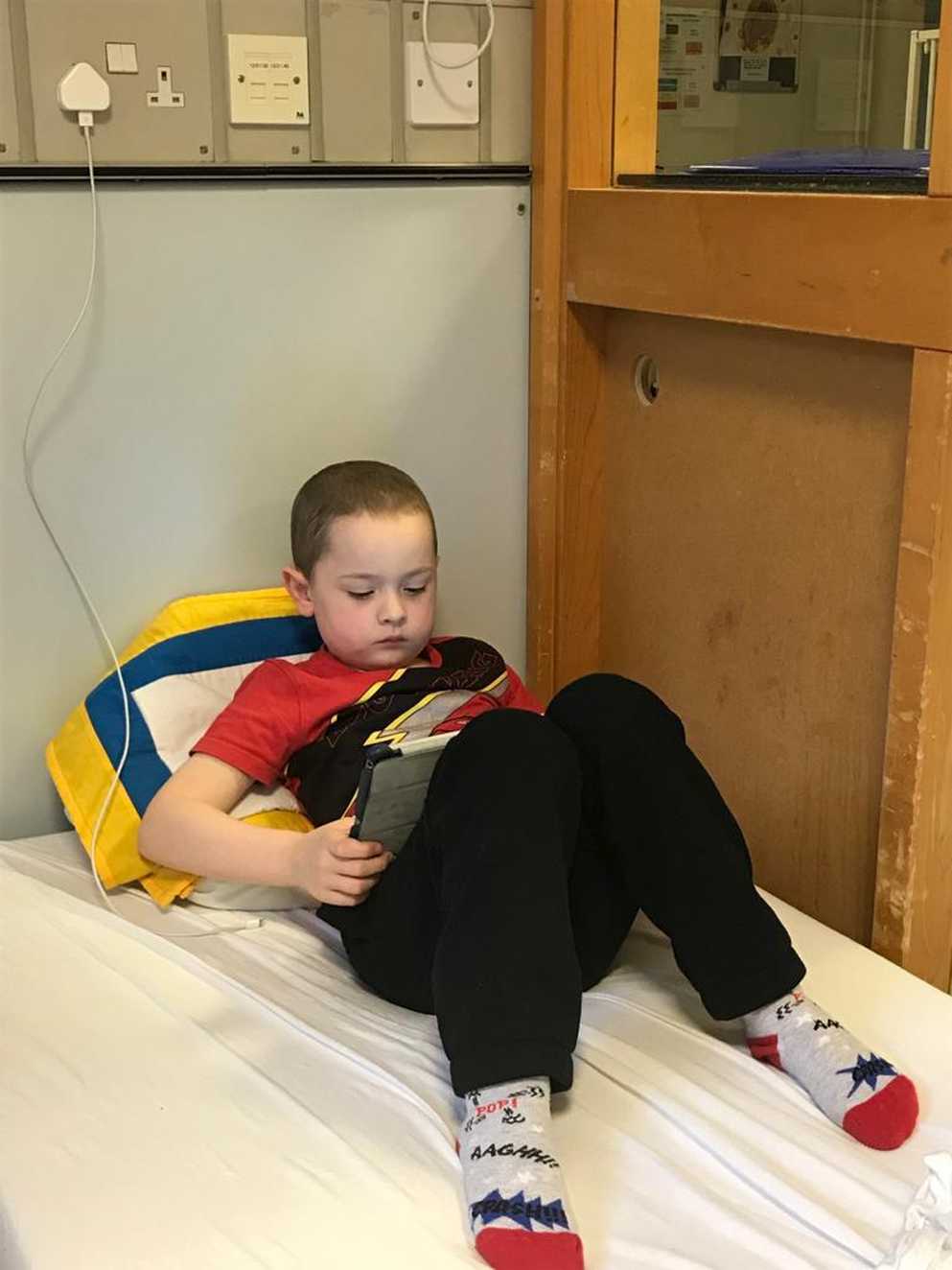 Ramsay as a young child is laying in a hospital bed watching his iPad. He is sat against a blue, yellow and white pillow.