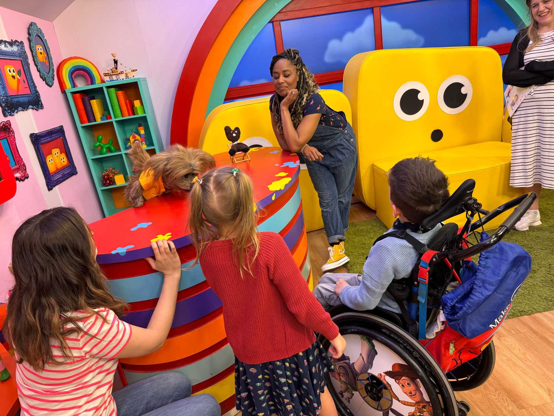 Dodge the Dog, a talking dog character from Dodge’s Pup School is behind a colourful desk, wearing a bright orange vest. Charlie sits in his wheelchair beside his sister, both saying hello to Dodge. Two CBeebies presenters stand with them.