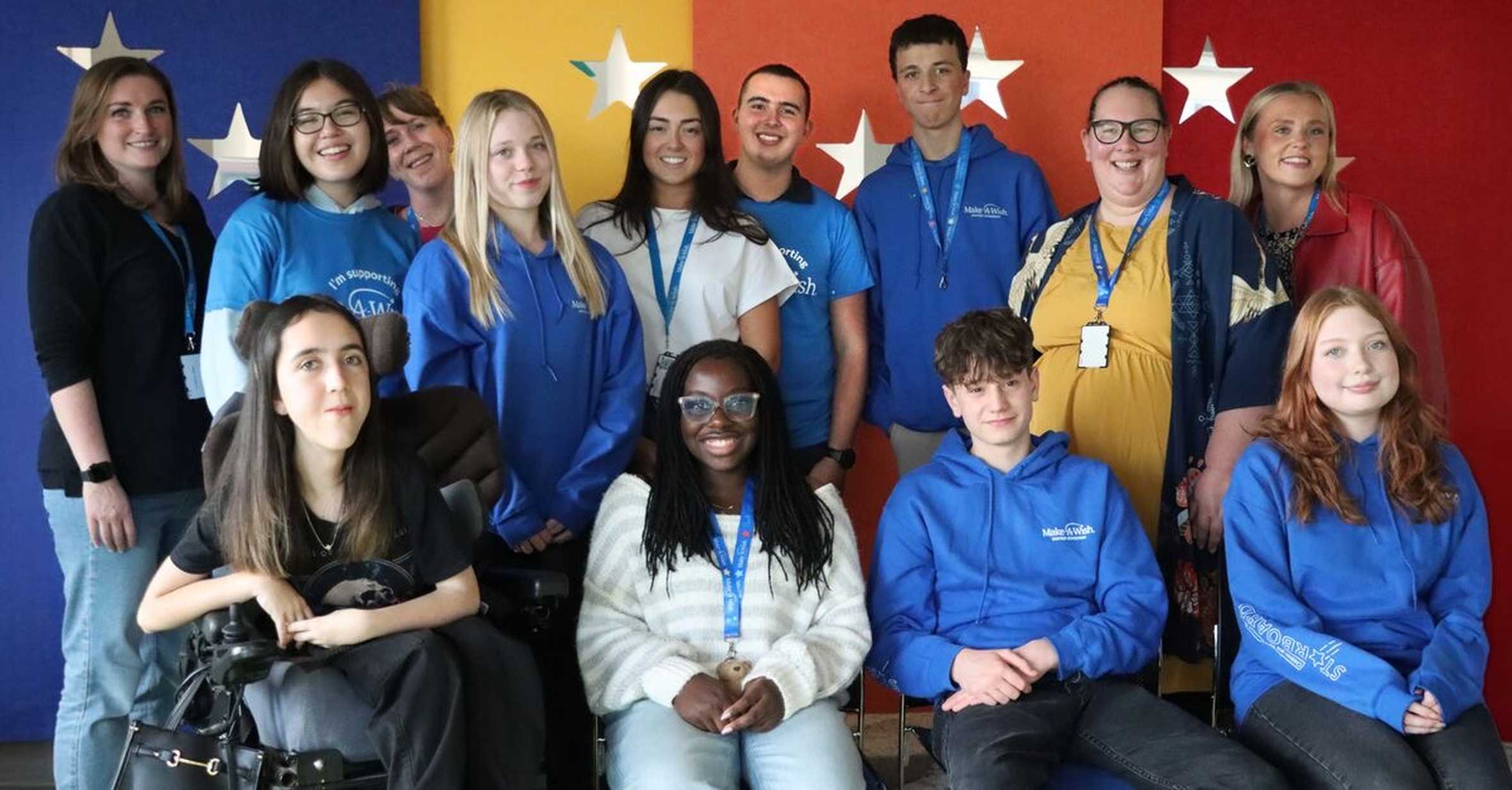 A group of young people are stood and seated smiling at the camera.  They are posing in front of a star and multi-coloured backdrop.