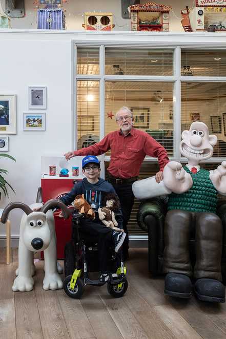 Mert is smiling in his wheelchair, wearing a blue Make-A-Wish UK cap. To his right is a large model of Gromit the dog, and to his left stands Aardman co-founder Peter Lord, also smiling. Beside Peter is a life-sized model of Wallace
