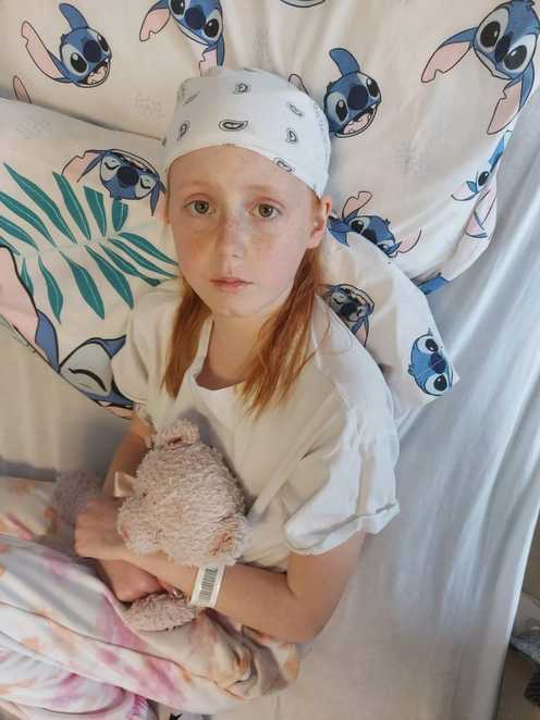 Faith sits in a hospital bed looking up. She is holding a soft toy and has bandages around her head.