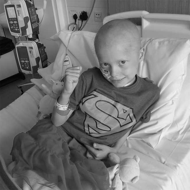 Black & white image of Indeg sitting up in her hospital bed, wearing Supergirl pyjamas.