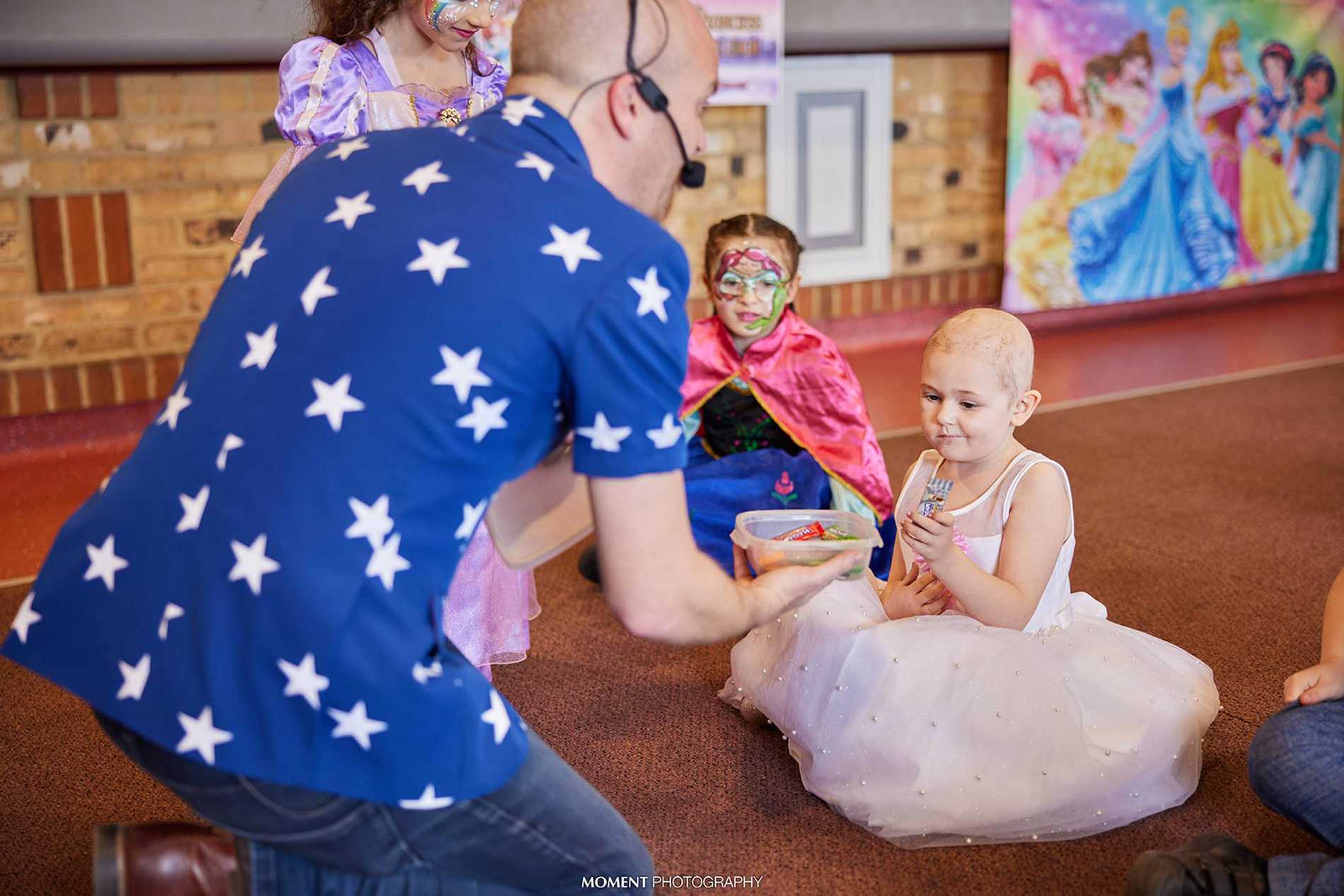 Ryleigh sitting on the floor while the entertainer, brightly dressed in a blue shirt with white stars, lets her choose something from a box.