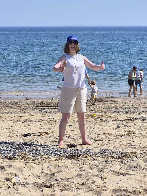 Millie smiles and gives a double thumbs up on the beach. She is wearing sunglasses and a Make-A-Wish hat