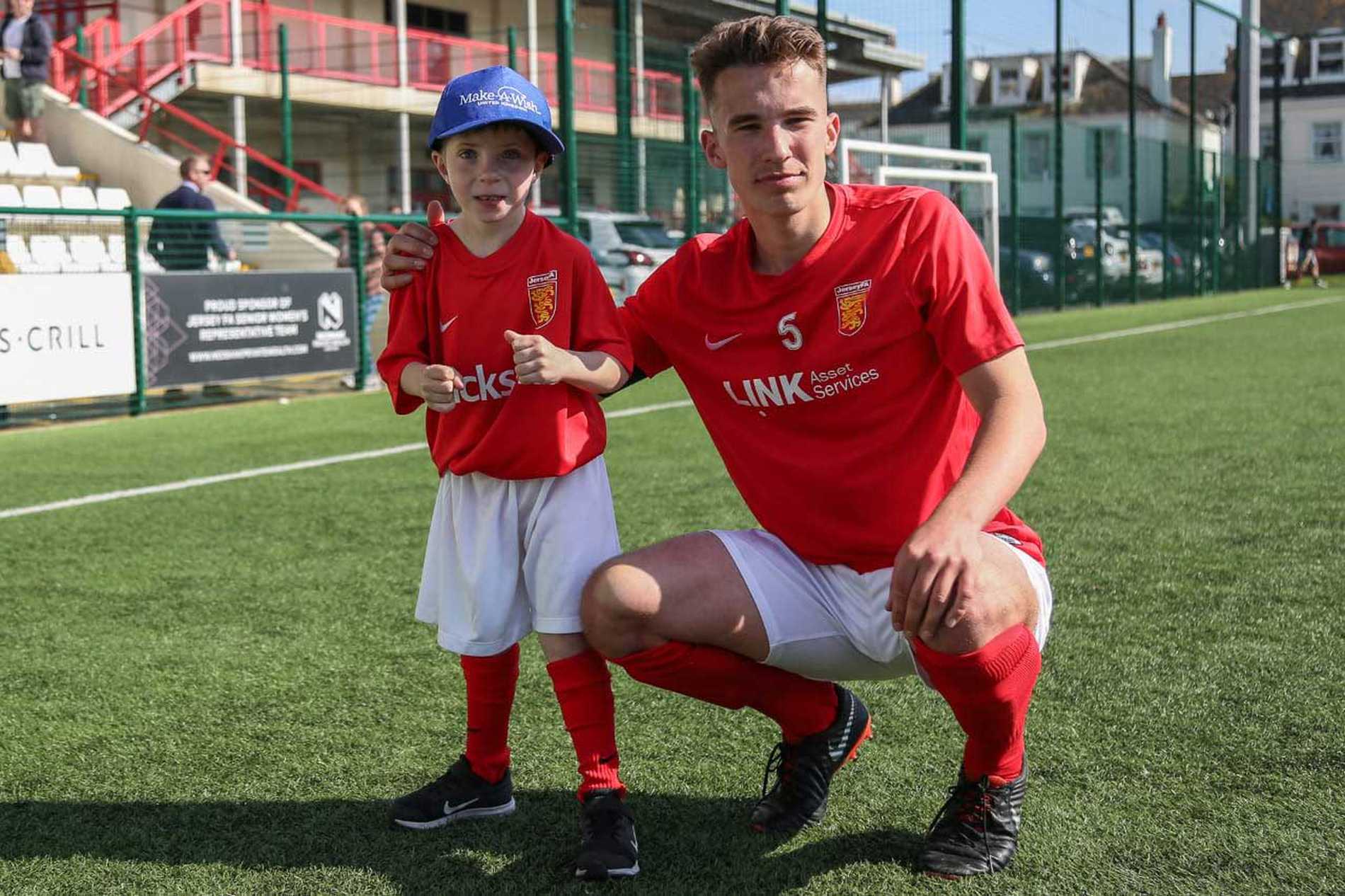 Wish child Jayden standing with a Jersey FC football player in their kits on the pitch
