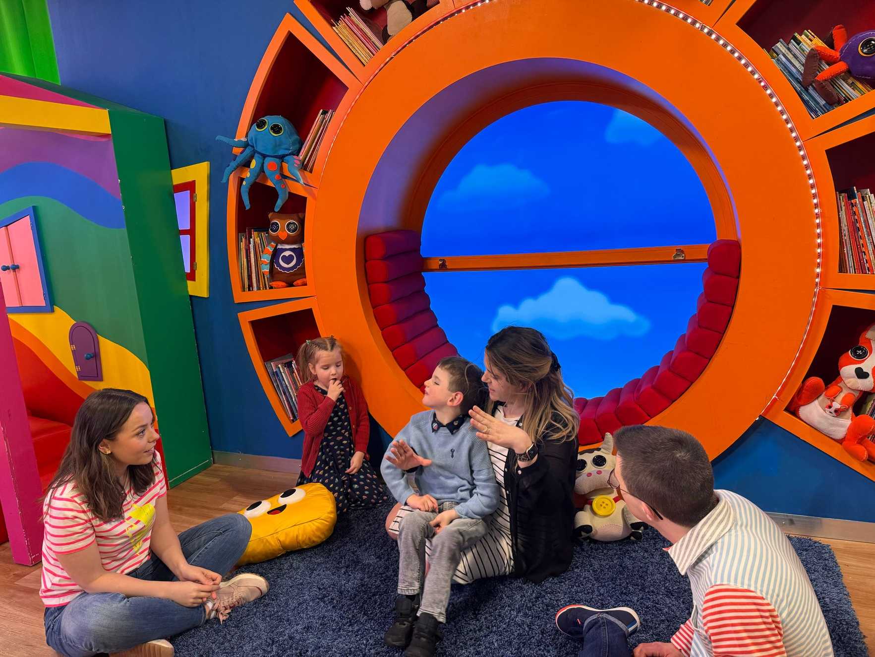 Charlie sits on the floor in front of a giant circular orange sofa, held by his mum. His sister is stood next to them, and they are talking to two of the CBeebies presenters.