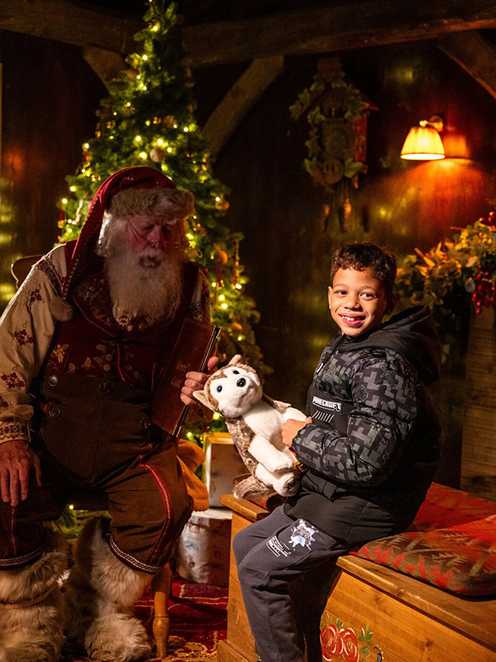 In a room full of Christmassy decorations, Kaiden smiles as he holds a grey and white Husky soft toy. Next to him is Father Christmas, holing a book which says ‘Good List’.