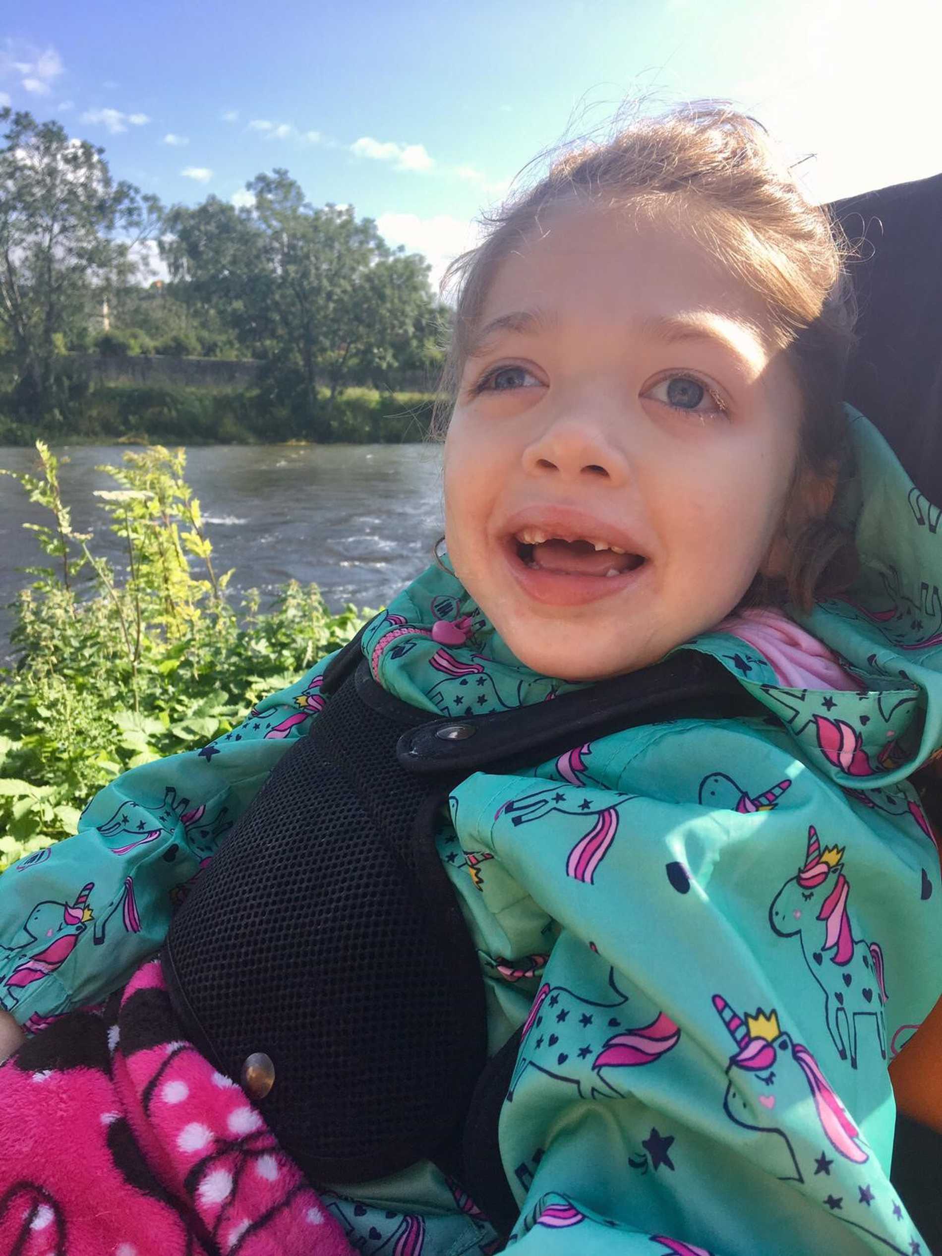 A close up of a smiling Aubree sitting in her wheelchair by a lake.