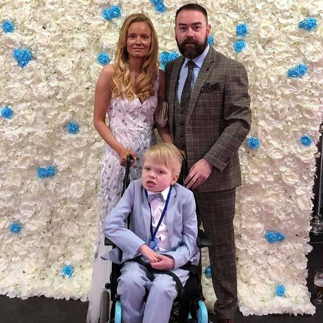 Jack posing in front of a floral wall with his mum and dad, during his prom.