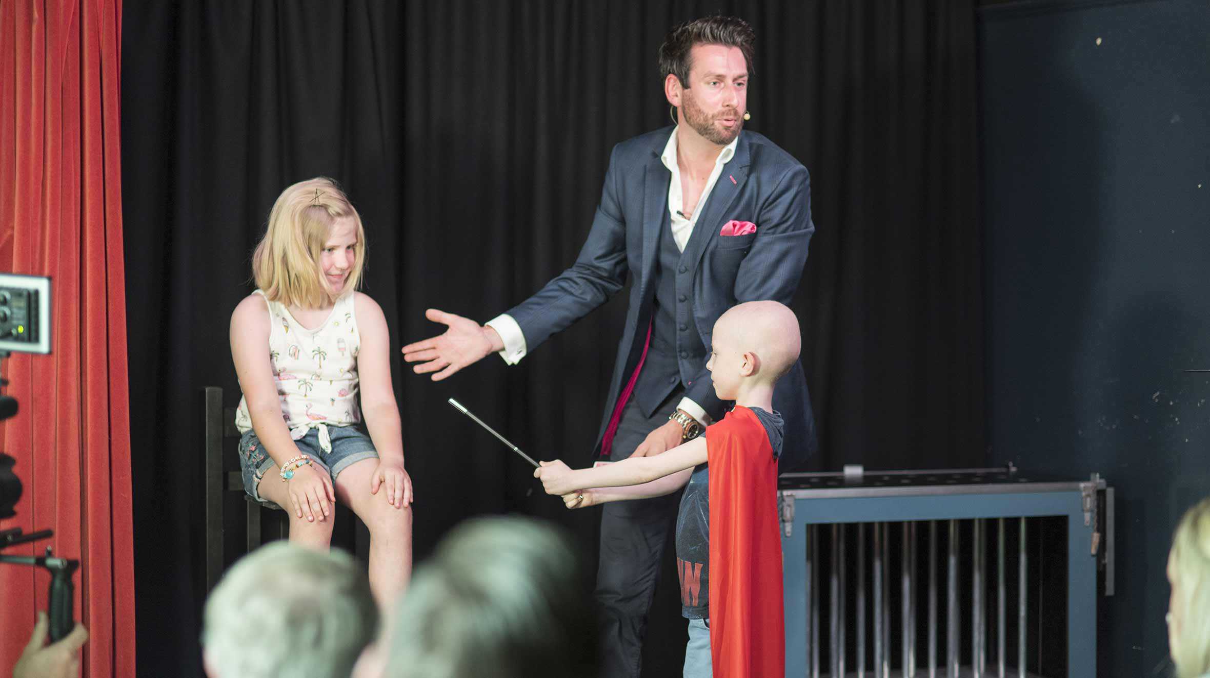 Madison sitting on a stool while Dexter waves a wand at her as Jamie looks on