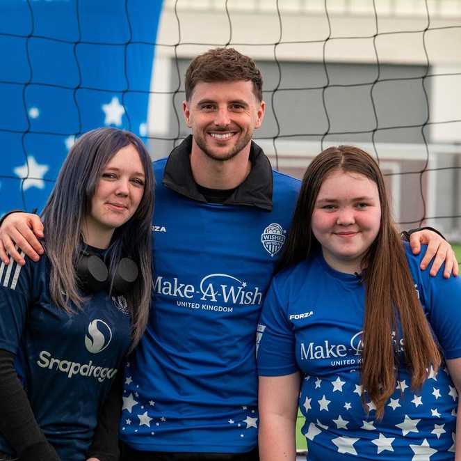 Robyn and her sister Miley stand either side of the footballer Mason Mount, who is smiling at the camera.  Mason and Robyn are wearing Make-A-Wish Wishes United football tops.