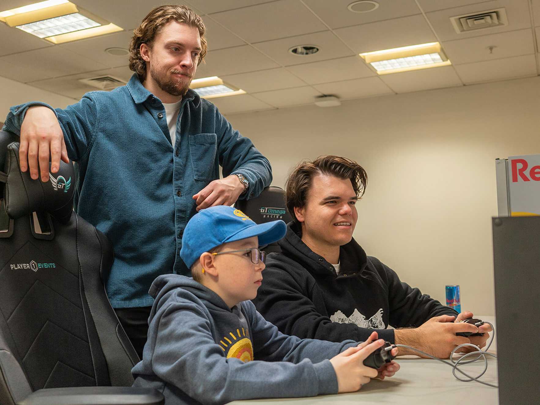 Ewan, wearing a cap, plays a computer game with Jelly, while Slogo stands behind them resting his arms on their gaming chairs