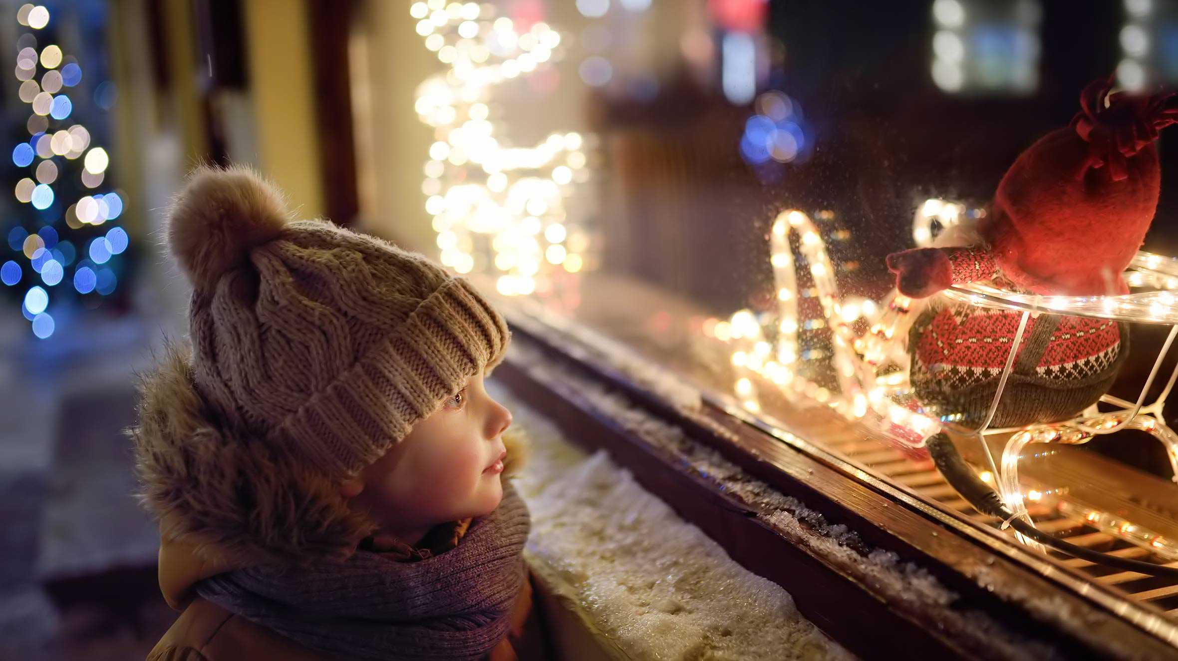 A child in a woolly hat and scarf, looking through a window at some Christmas lights.