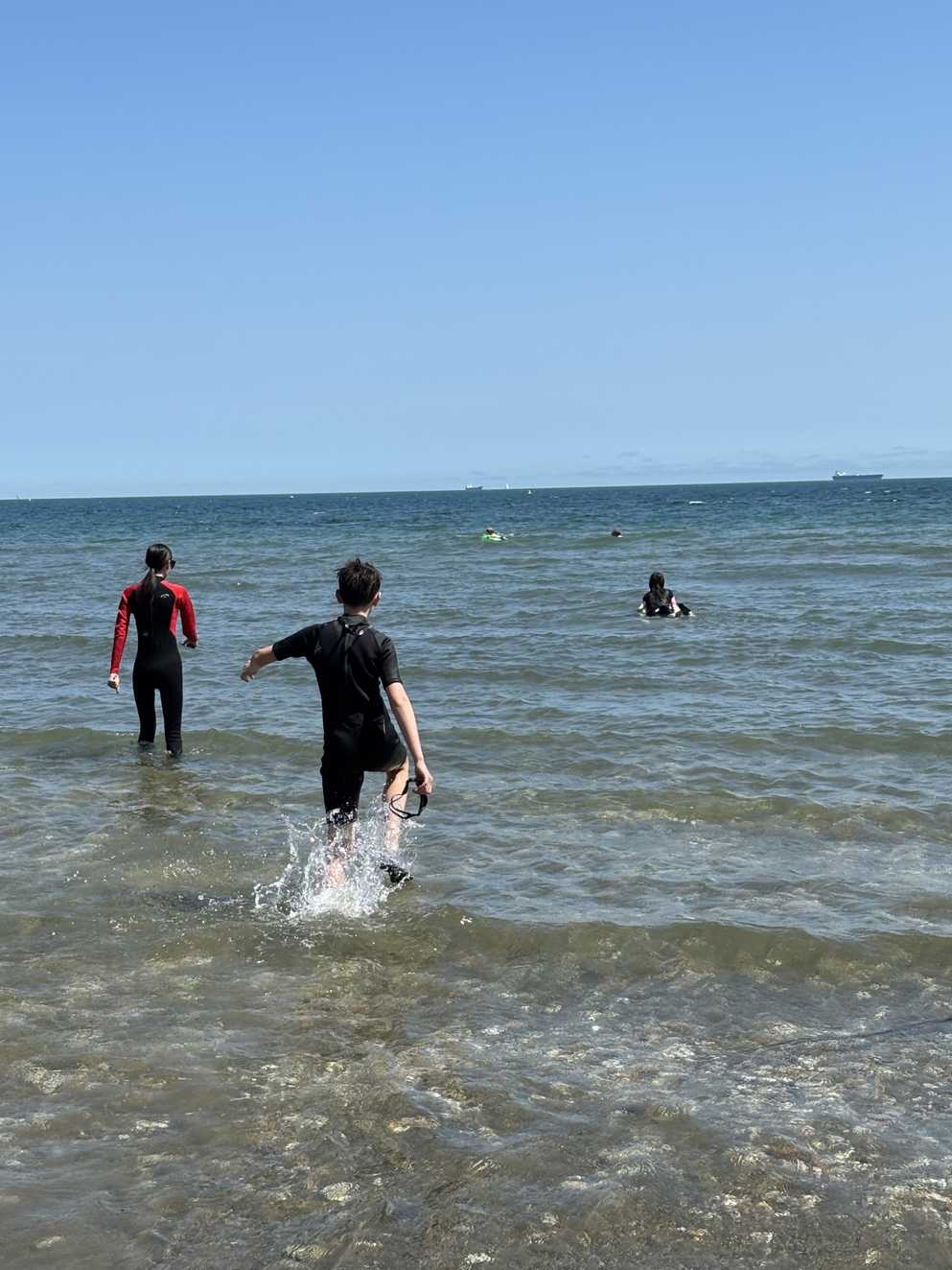 Ramsay and his sisters walk out to sea in their wetsuits and goggles. The sea and the sky are blue as Ramsay’s feet splash through the water.