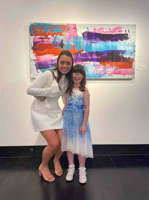 Two people stand smiling in front of a colourful abstract painting in a gallery. One person wears a white outfit and the other wears a blue and white dress with sneakers.