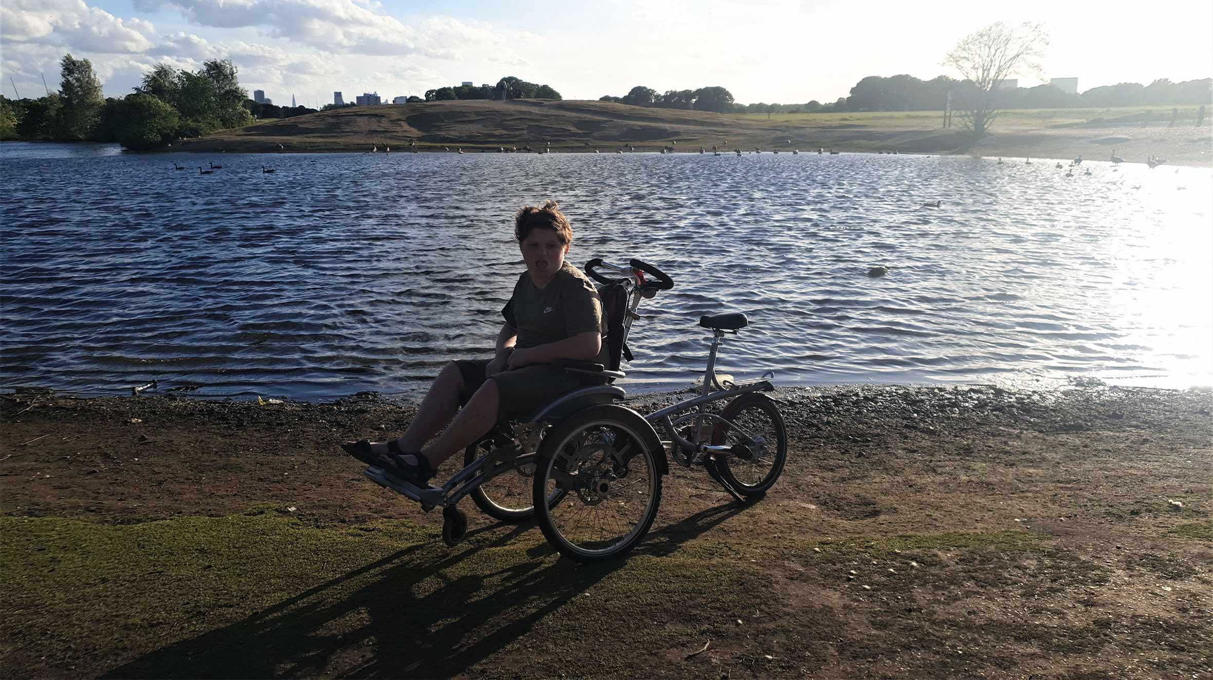 A picturesque view of Eric sitting on his bike by a lake in the sunshine, with the London skyline behind him.