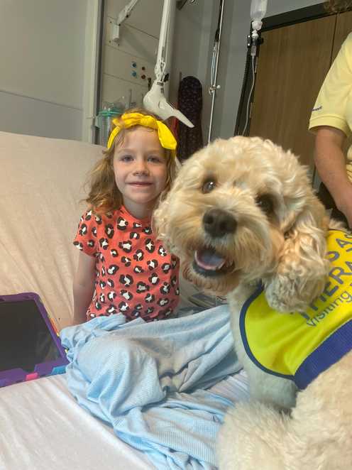 Ella sits smiling on a hospital bed while undergoing treatment, with a therapy dog on the bed with her