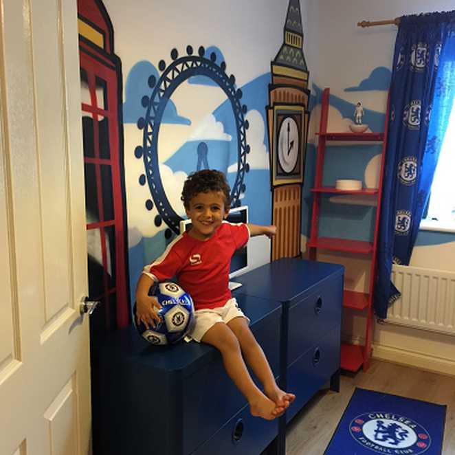 Wish Child Mohammad sitting on his set of drawers in his new bedroom, holding on to a football and smiling at the camera