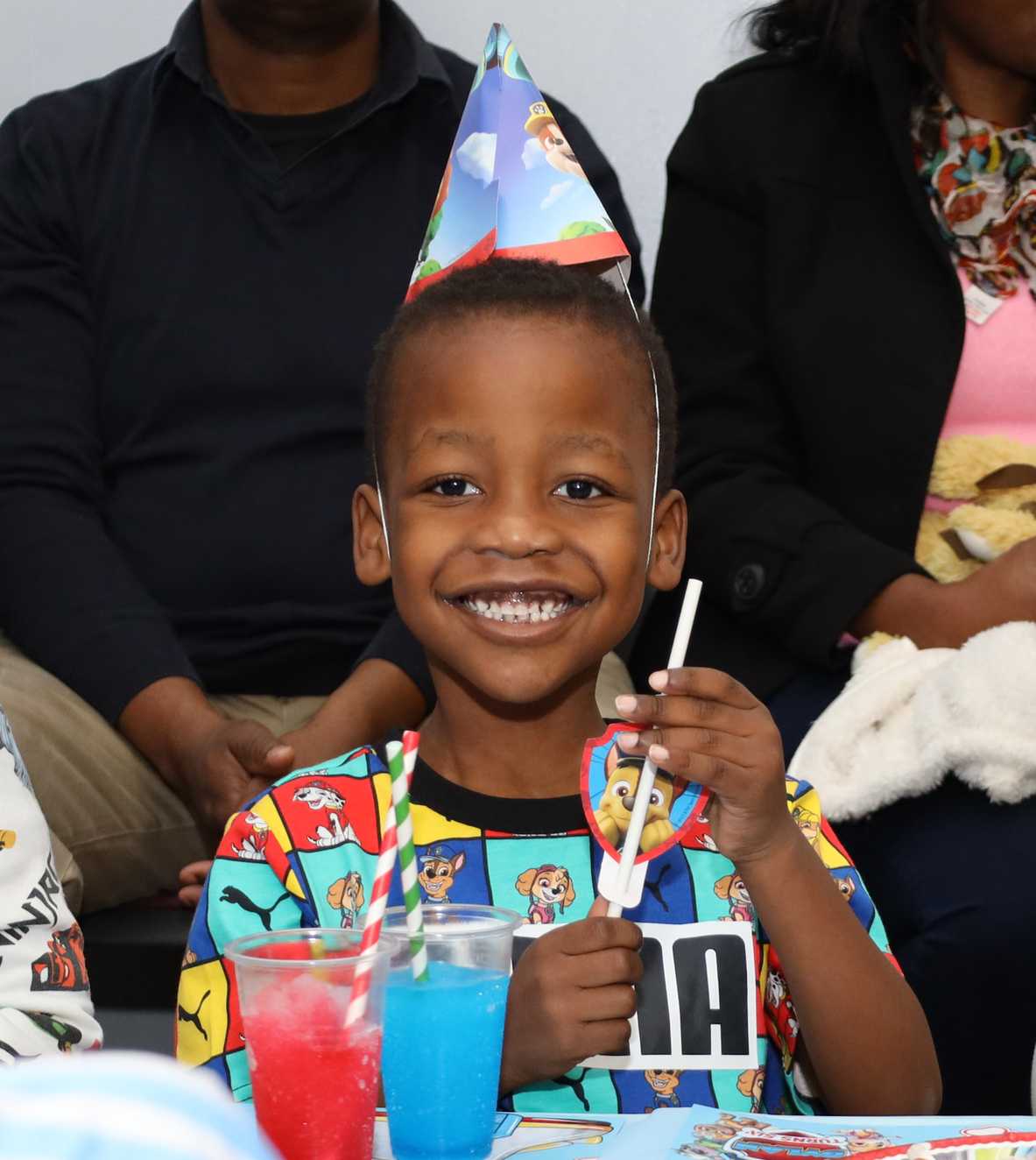 Dylan has a large toothy smile on his face and is looking straight on at the camera. He is wearing a brightly coloured t-shirt, Paw Patrol party hat and holding a Paw Patrol straw. There are red and blue frozen slushie drinks in front of him on the table.