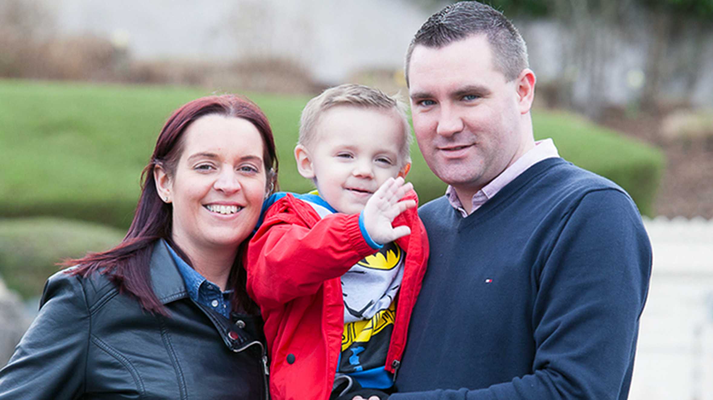 Zak, wearing a red jacket, being held by his mum and dad.