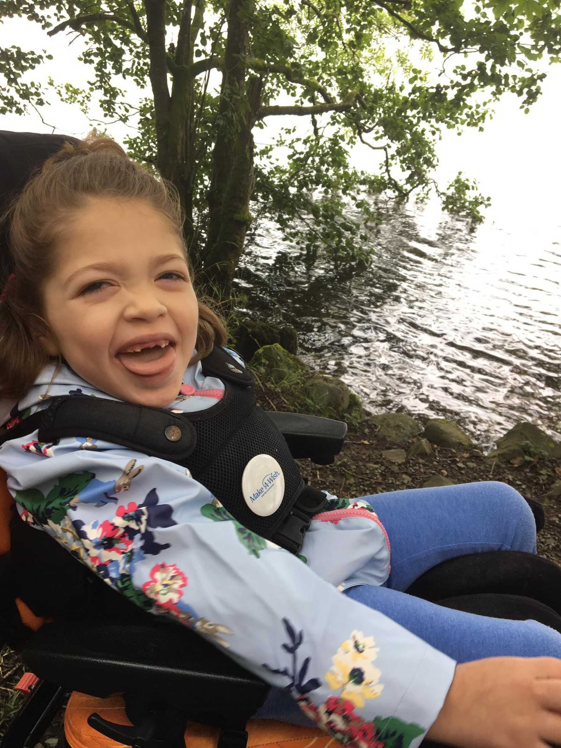 A close up of a smiling Aubree sitting in her wheelchair by a lake.