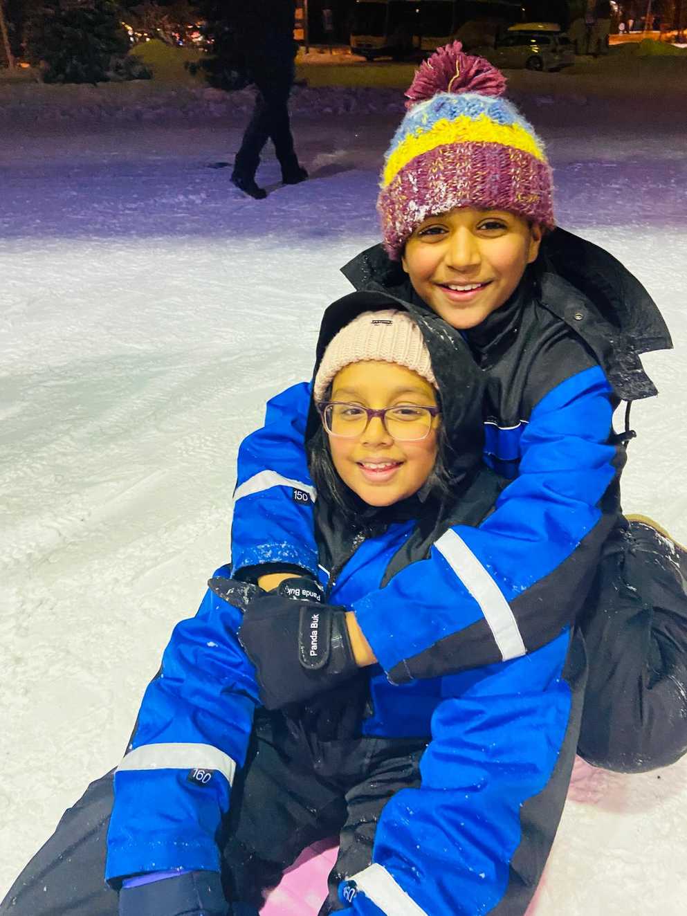 Zara is sitting on a sledge, with her brother hugging her from behind. Both are smiling widely, wearing woolly hats and black and blue snowsuits. The snowy ground is visible in the background.