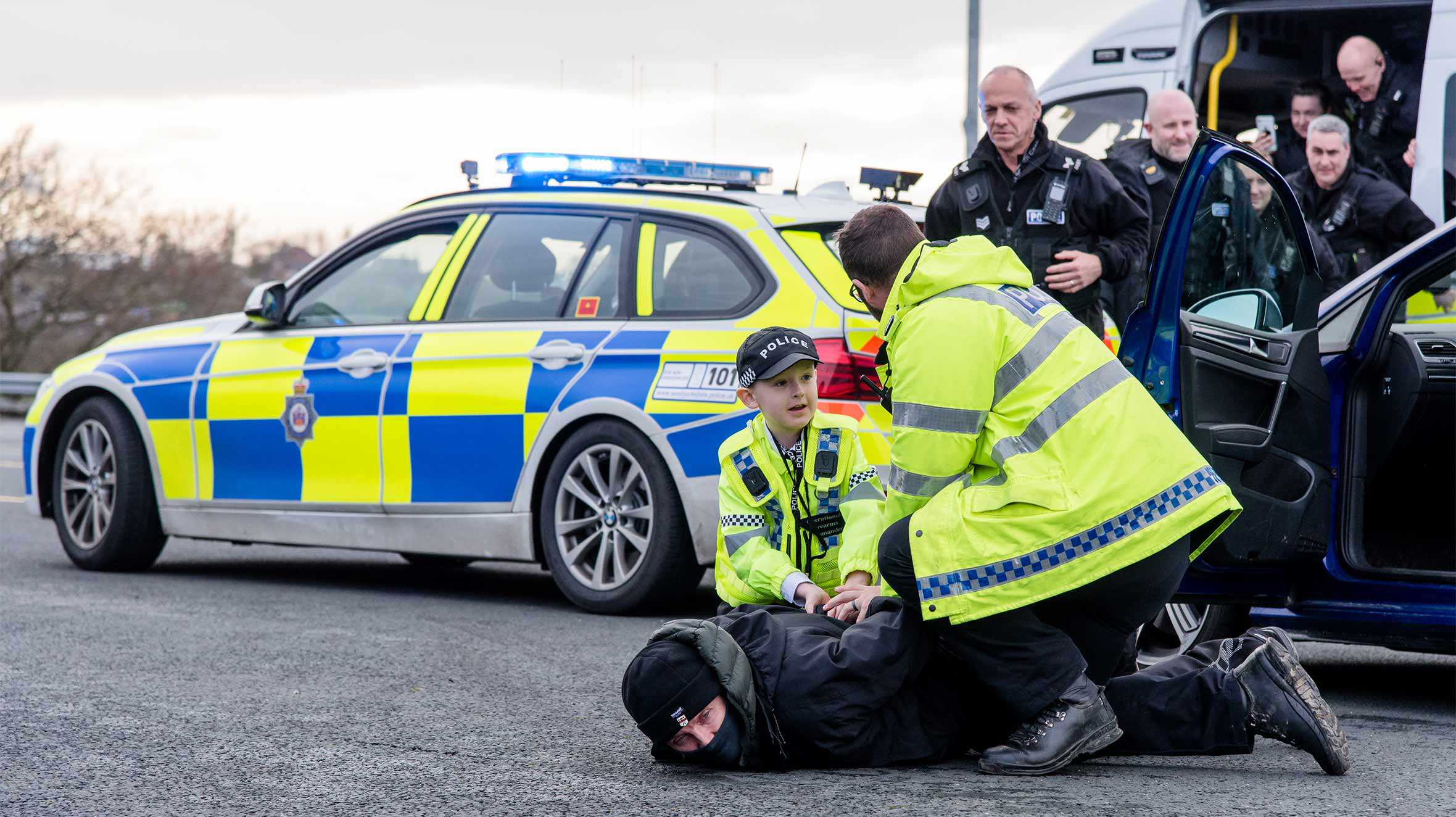 Allan and another officer subduing a suspect on the floor, with a police car and riot van in the background.