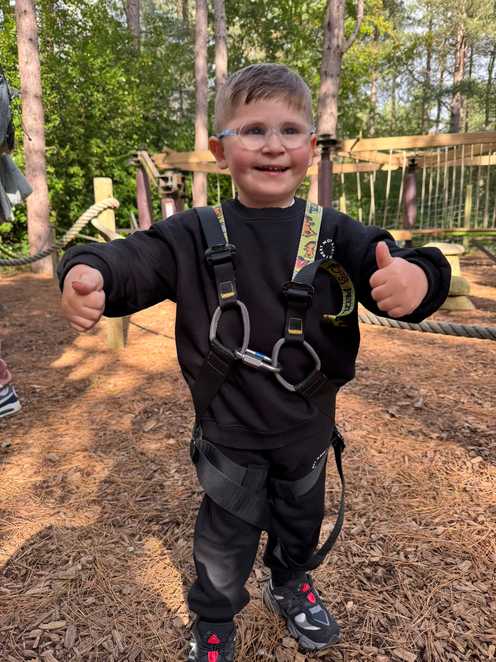 Dzemil is smiling with his thumbs up standing in the woodlands at Centre Parcs. He has a harness around him and some wooden obstacles behind him.