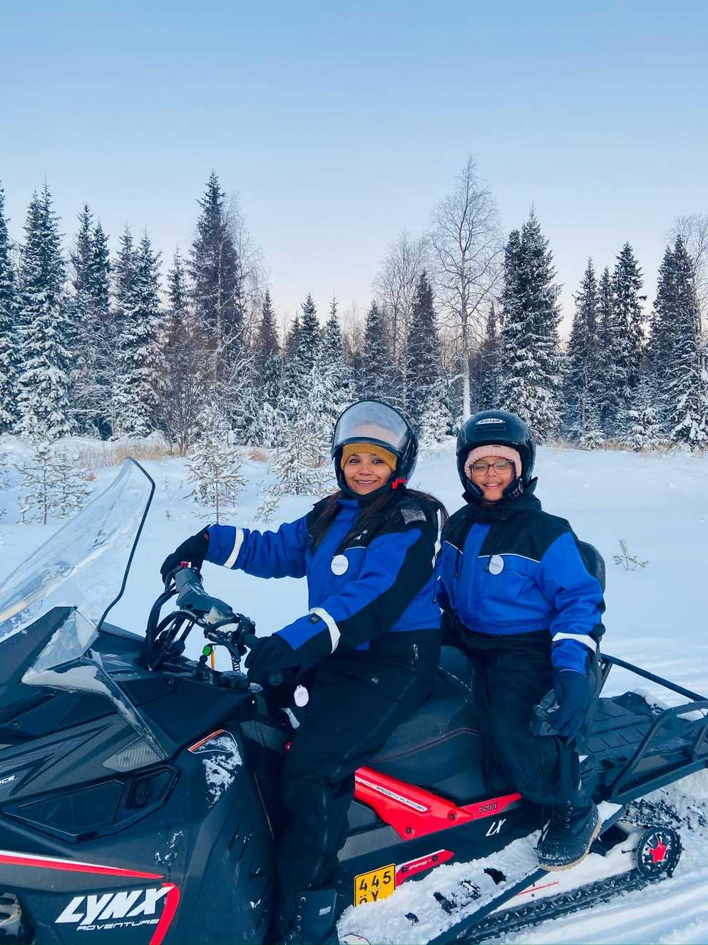 Mum and Zara are sitting on a black and red snowmobile, both dressed in black and blue snowsuits, with snow lined trees in the background. They're wearing helmets with woolly hats underneath. Zara’s mum is holding the handlebars, and they are smiling.
