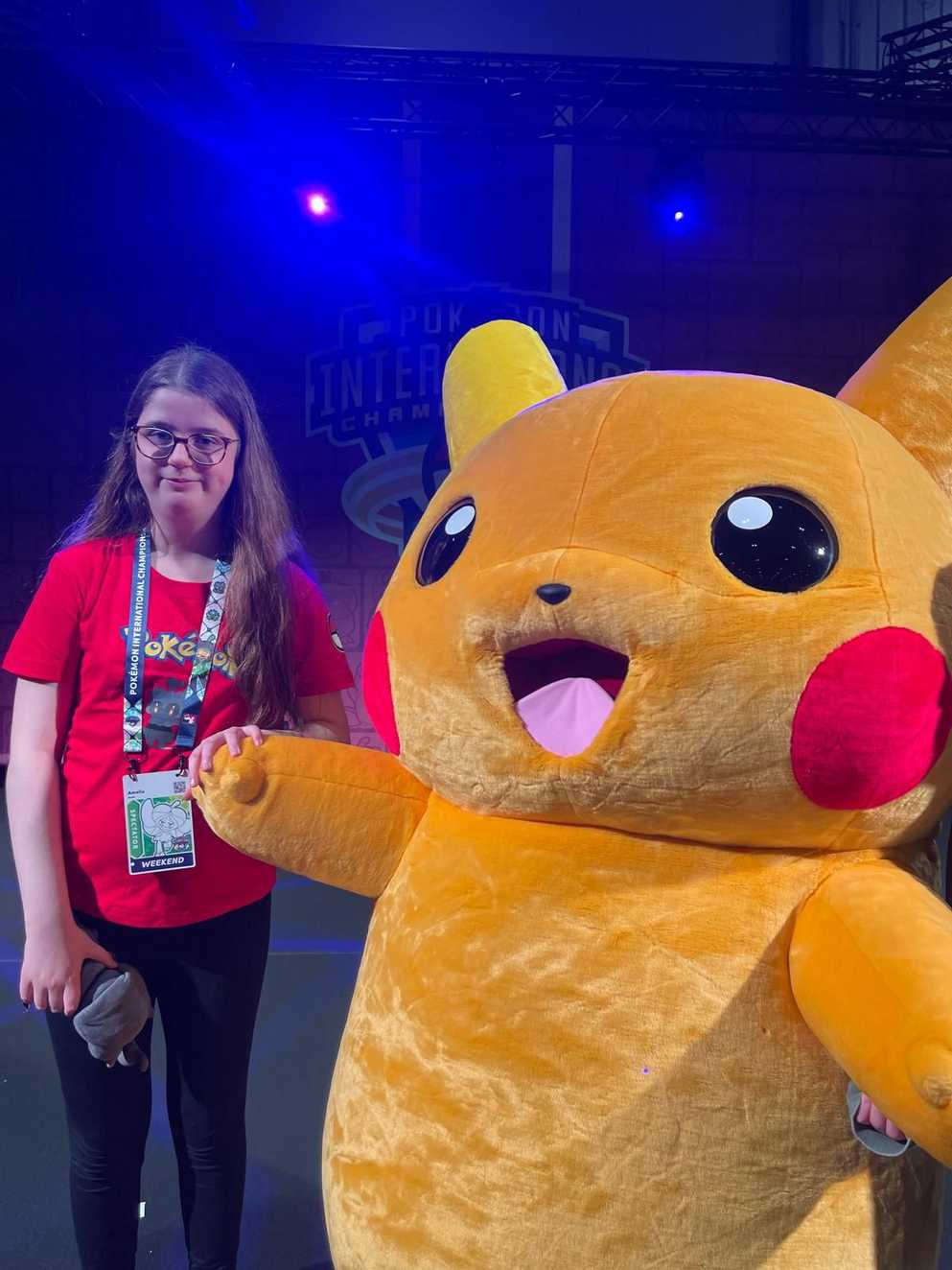 Amelia is wearing a red Pokémon t-shirt and dark skinny jeans. Amelia is on a stage lit with blue light and is standing next to a large Pikachu mascot at the Pokémon Championships in London. Both are smiling widely for the camera.