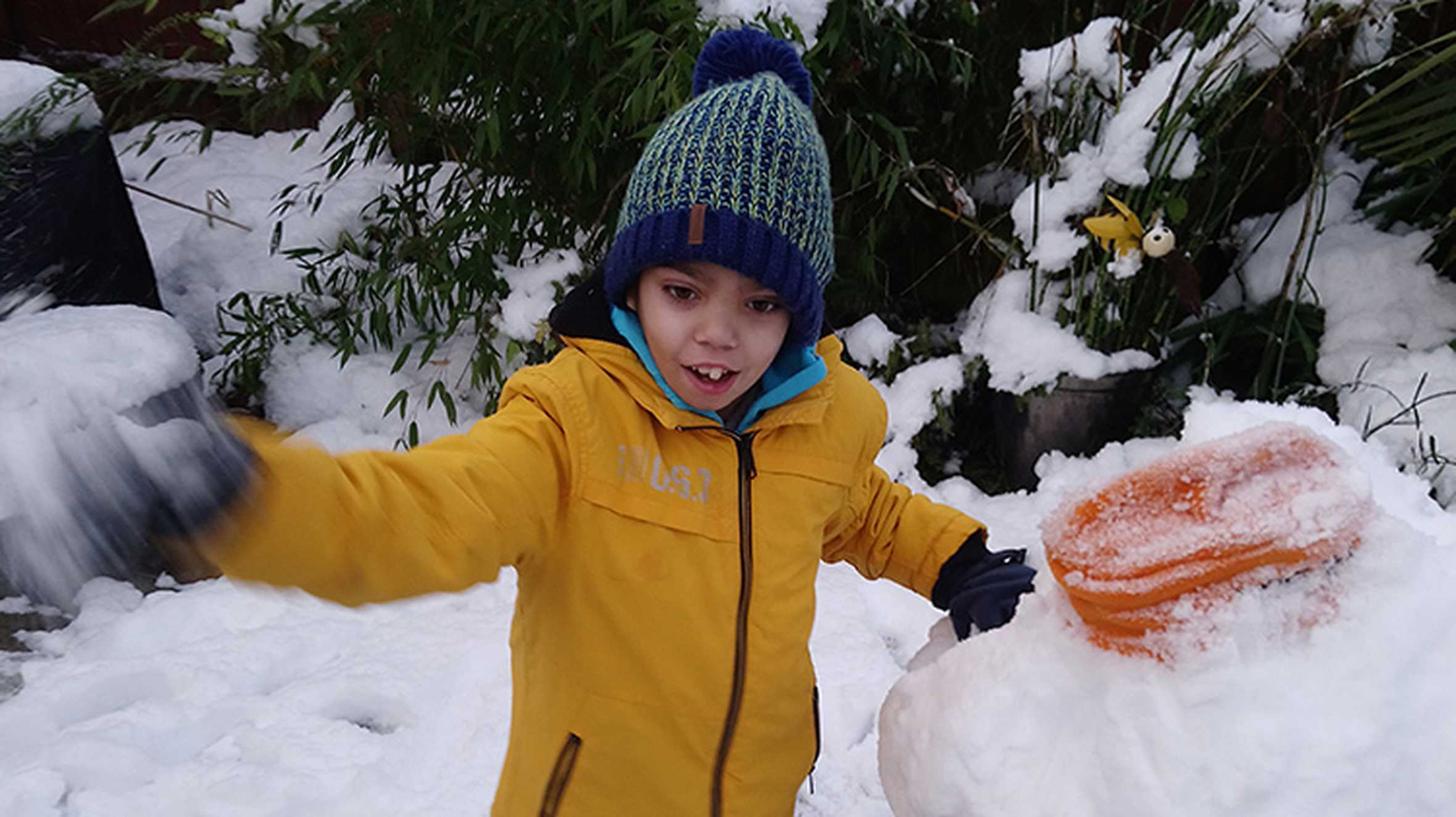 Zaid playing in the snow, dressed in a yellow jacket and blue bobble hat.