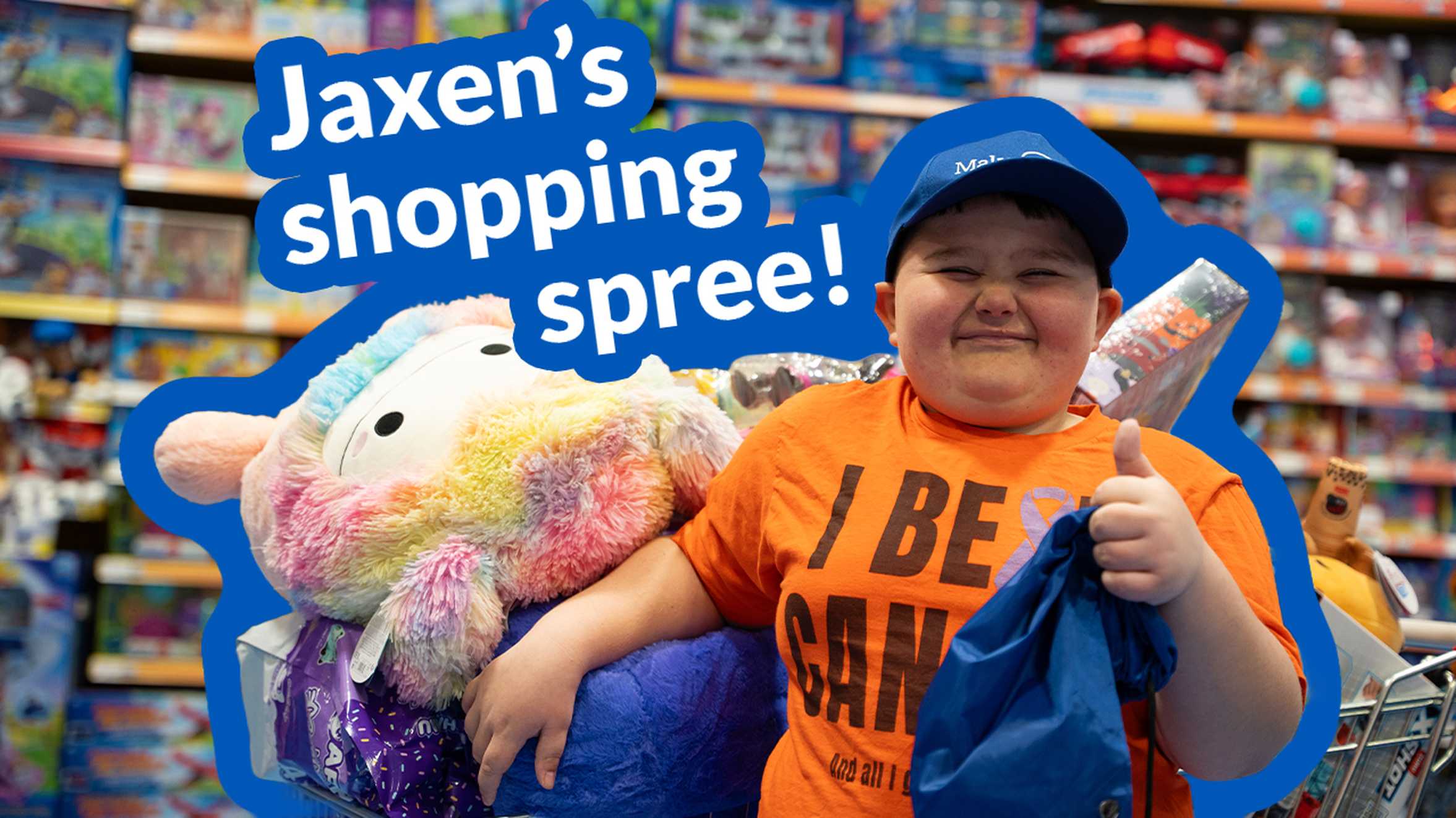 Jaxen on his shopping spree, smiling and giving a thumbs up to camera.  Stood in front of a shopping trolley filled with toys.  There is text on the picture "Jaxen's shopping spree"