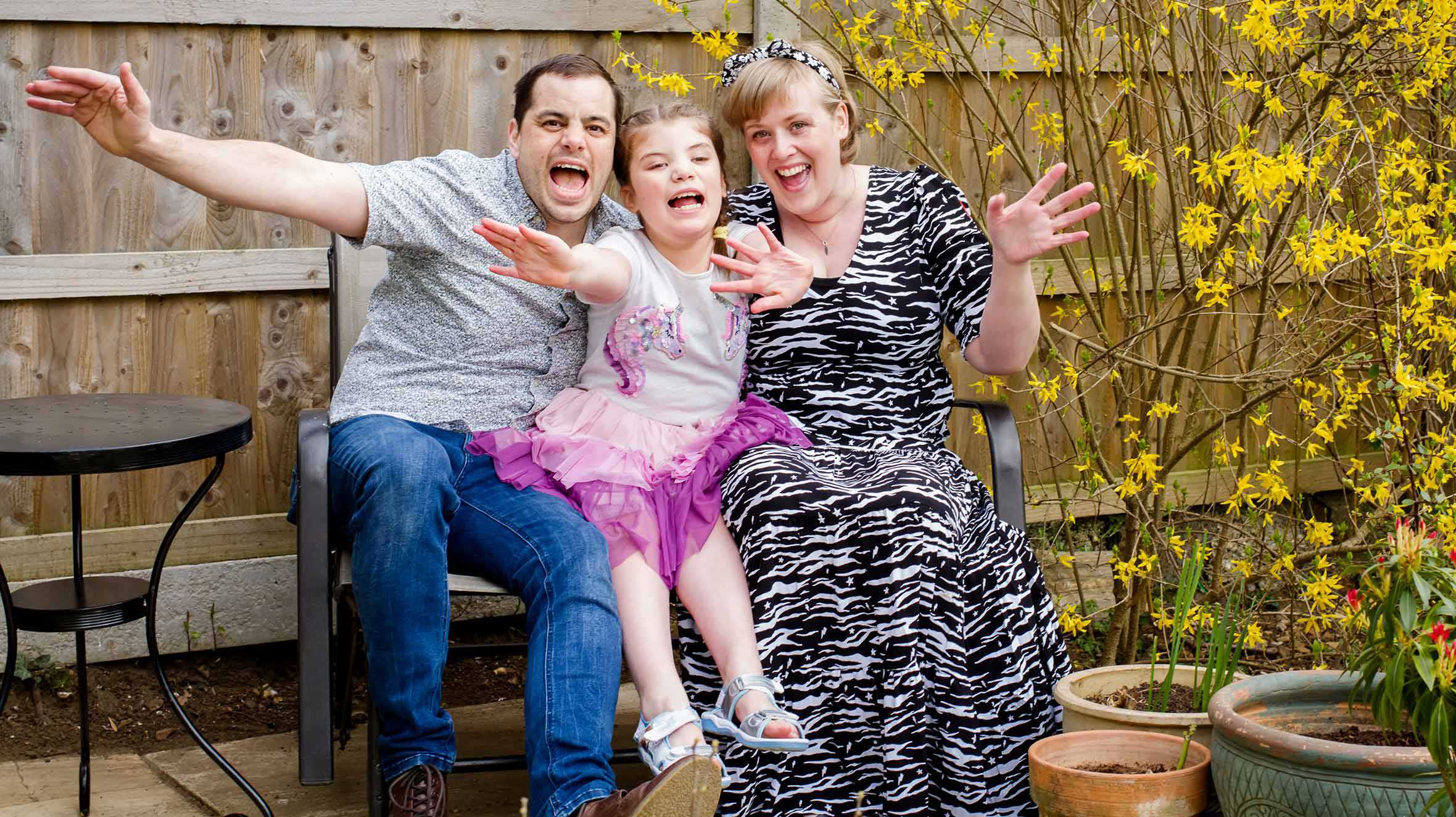 Gracie and her parents on their garden bench, smiling and waving at the camera