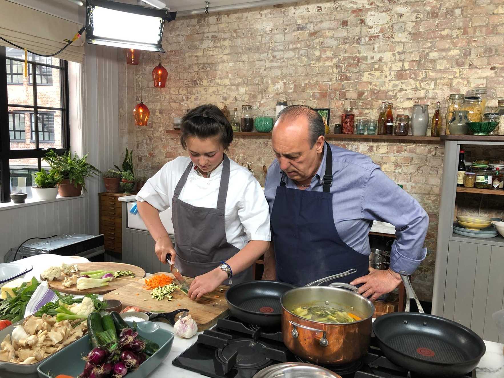 Sara Millar and chef Gennaro Contaldo are in a kitchen setting with exposed brick walls. Sara chops vegetables on a cutting board, while Gennaro observes. The stove has a pot and pan, with visible ingredients like garlic and fresh vegetables nearby.