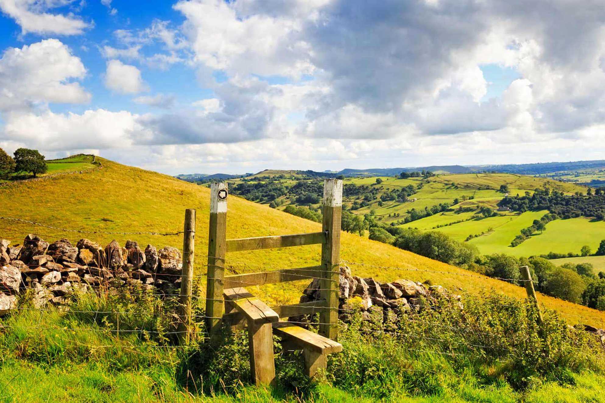 A scenic view of a stile over a dry stone wall with the Peak District countryside stretching of into the distance.