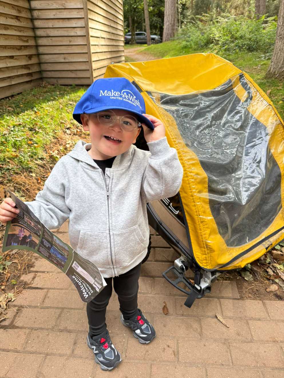 Dzemil stands next to a bright yellow trailer, which can be attached to a bike. Dzemil is smiling wearing a dark blue Make-A-Wish UK hat, holding a paper map of Centre Parcs.