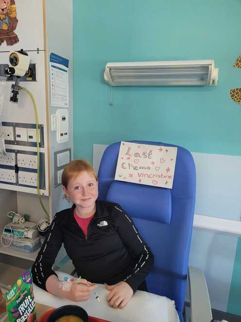 Faith is sitting in a blue hospital chair, which has a paper sign on it with black and pink writing which says “Last chemo vincristine.” Drawn around the sign are hearts and stars. Faith is smiling with a medical line in her hand