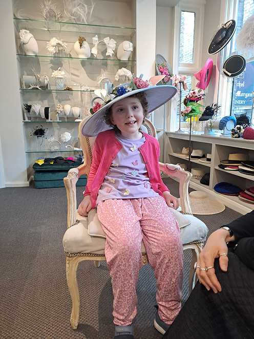 Penelope is wearing a lilac hat inside Rachel Trevor-Morgan Millinery. The hat has light blue ribbons, dark blue, white and pink flowers, and green leaves.