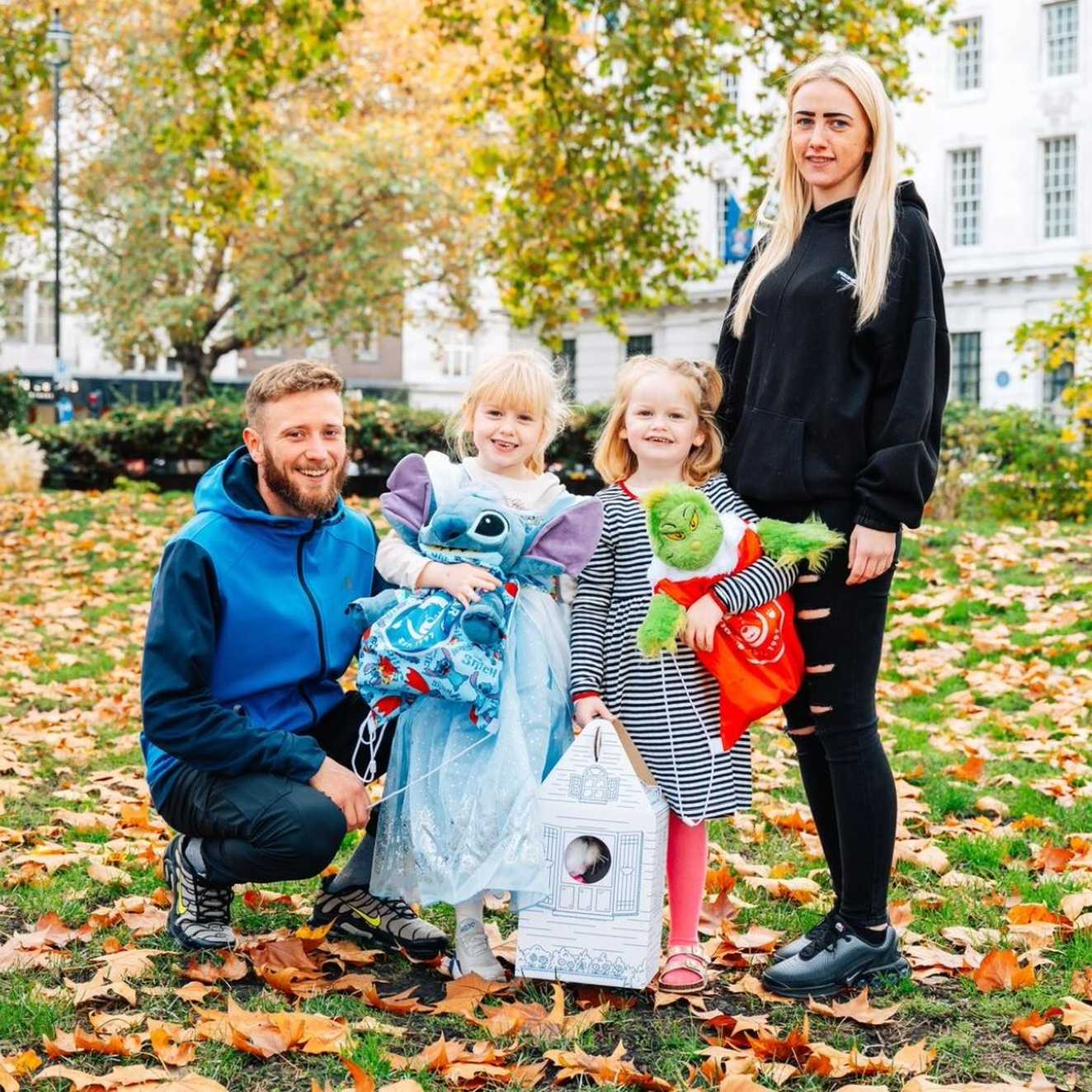 Demi stands next to her mum, twin sister and dad. Demi and her sister are holding toys including a green The Grinch toy, and a Blue Stitch toy. They’re all standing in a green space in London, with leaves surrounding them.
