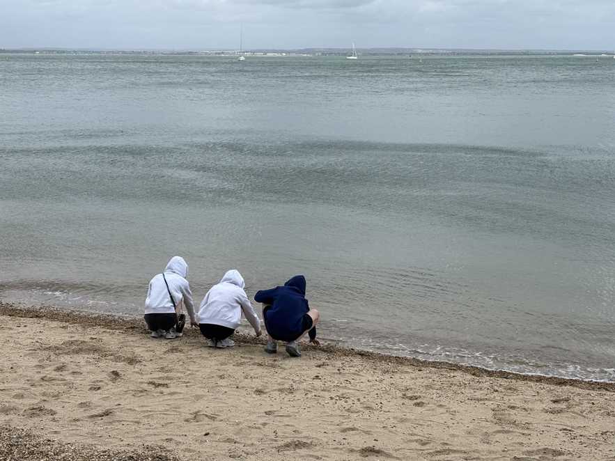 Ramsay and his siters plan in the sand with their hoods up on a cold day. The sea is light blue and the sand is golden.