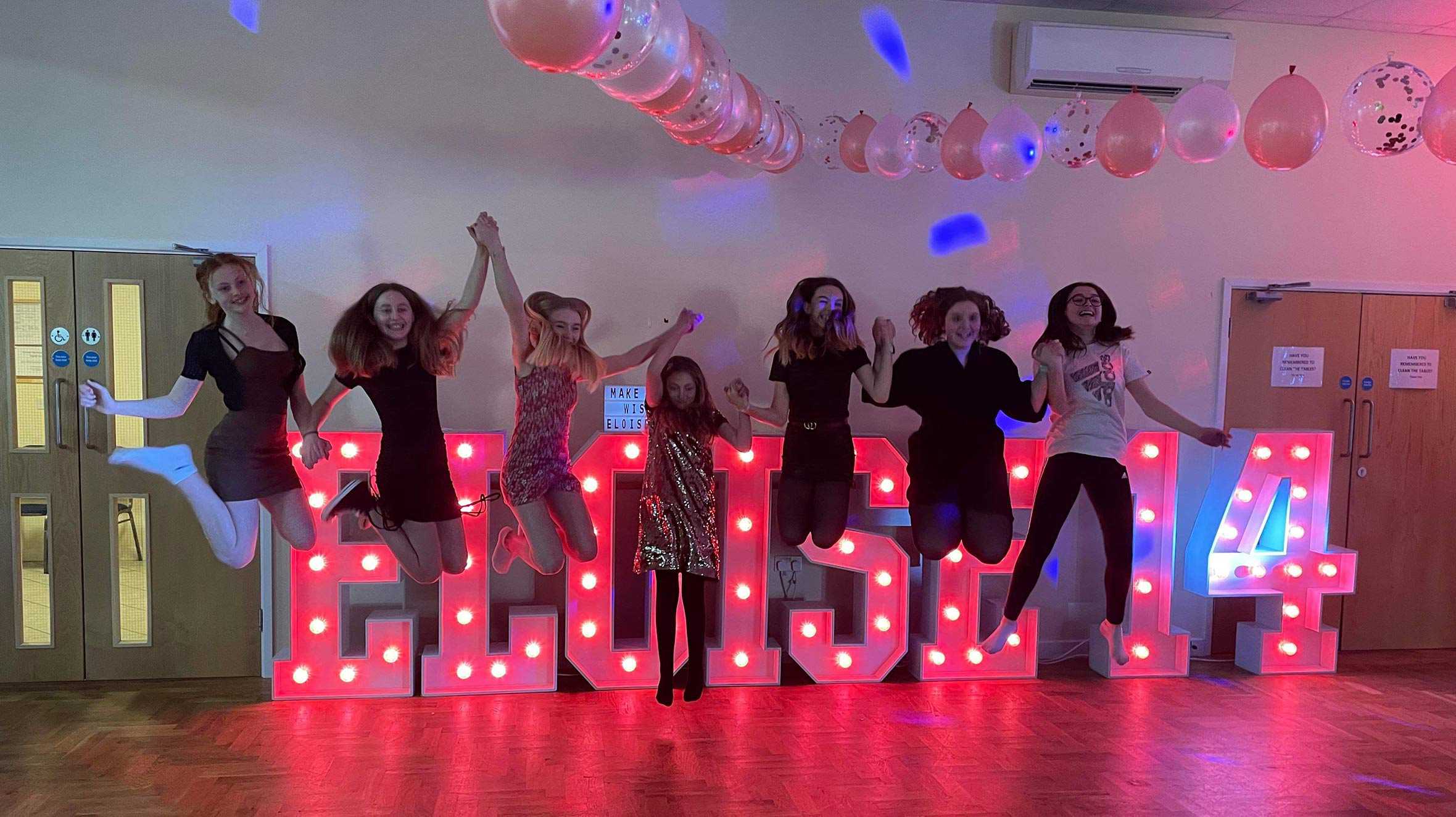 Eloise and her friends jumping in the air in front of her name, spelled out in large pink light up letters.
