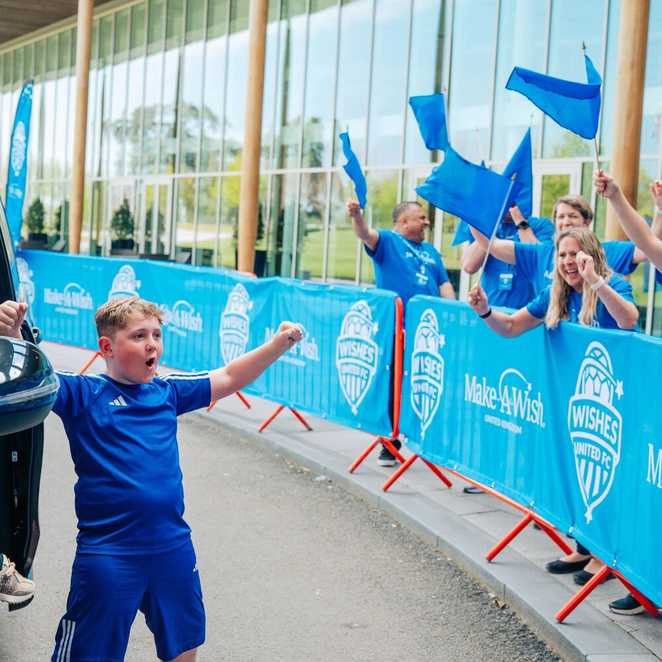 A boy is holding his arms aloft, his face filled with excitement as people cheer and wave flags from behind the barriers.  He has just got out of a taxi.